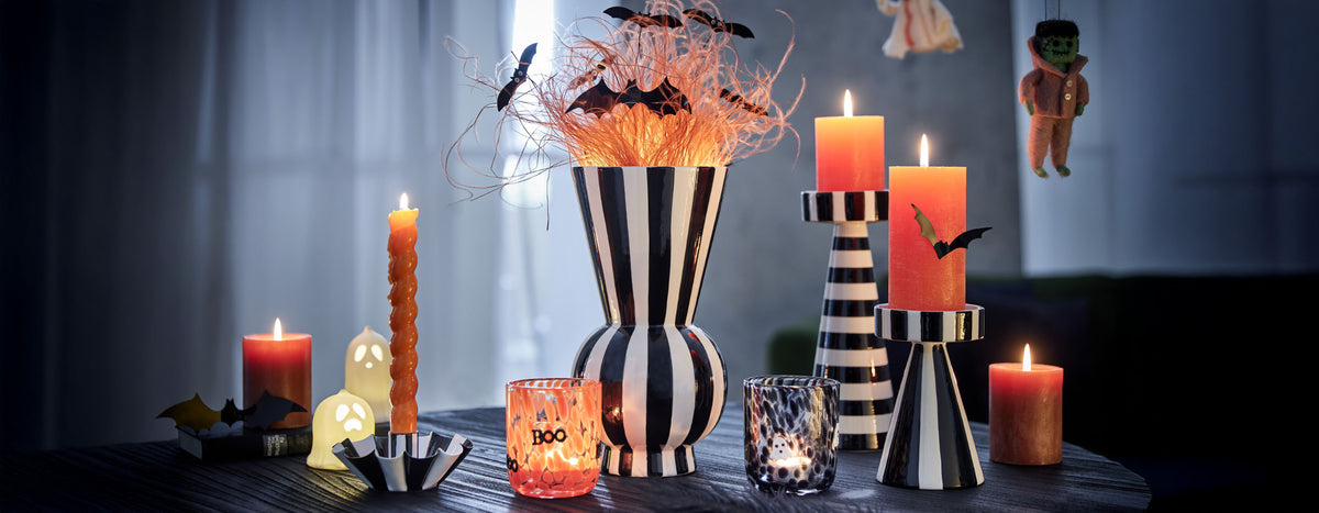 Halloween-Dekoration auf einem gedeckten Tisch mit orangefarbenen Kerzen, schwarz-weiß gestreiften Vasen und Kerzenhaltern, kleinen Geisterlichtern und Fledermäusen.