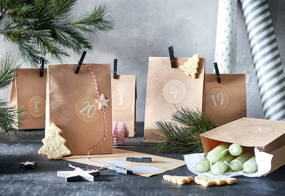 Adventskalender aus braunen Papiertüten mit Zahlen-Aufklebern, schwarzen Klammern, grünen Stabkerzen und Keksen auf dunklem Holztisch.