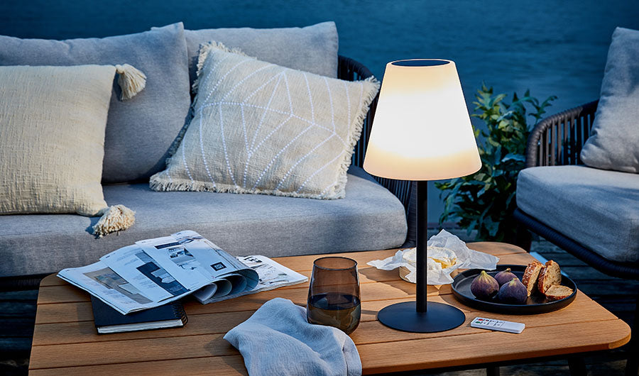 Leuchtende Außenlampe auf Holztisch neben grauer Gartenlounge am Abend