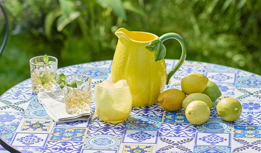 Gelber Zitronen-Keramikkrug auf blauem Mosaik-Gartentisch mit Trinkgläsern