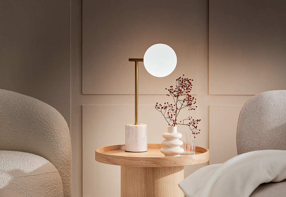 Moderne Designer-Tischlampe mit runder Glaskugel und Messing-Gestell auf einem runden Beistelltisch aus hellem Eichenholz vor Wandpaneelen.