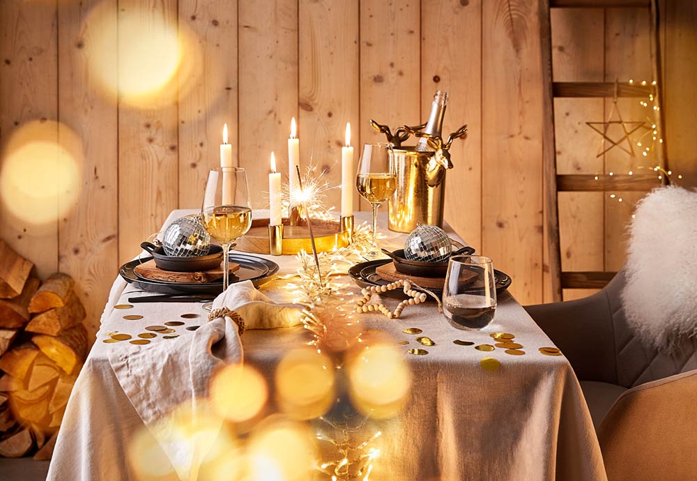 Festlich gedeckter Tisch mit Lichterkette, Kerzen, Weingläsern, Sektkühler in Elchform und Discokugeln vor einer Holzwand im Chalet-Stil.