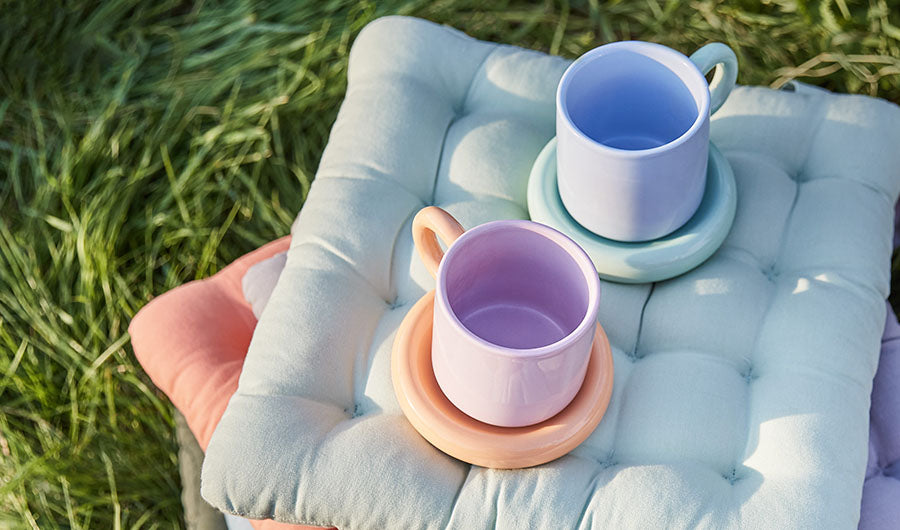 Zwei pastellfarbene Tassen auf blauem Sitzkissen im frischen Gras
