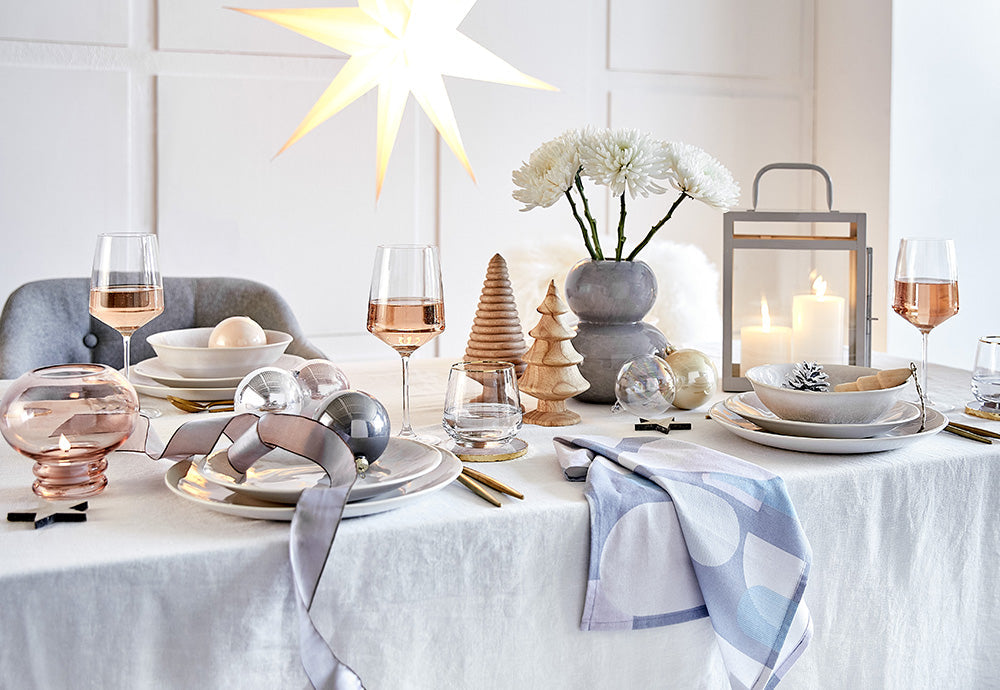 Festlich gedeckter Weihnachtstisch mit weißem Leinentuch, Weingläsern, Keramik-Geschirr, Holz-Deko-Bäumen und leuchtendem Stern im Scandi-Stil.