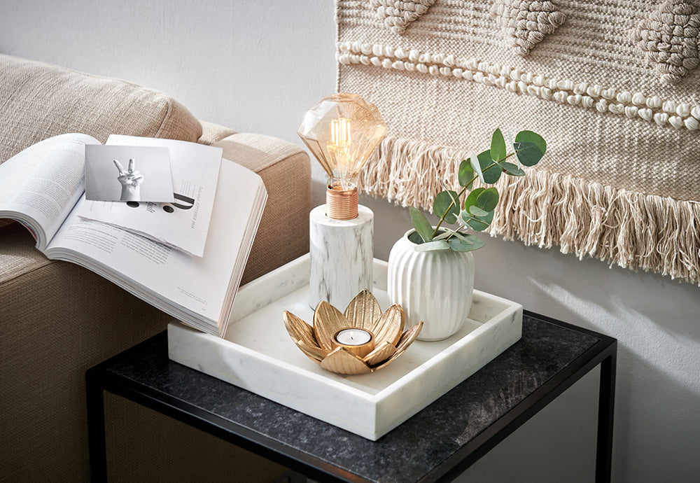 Weißes Marmor-Tablett auf schwarzem Beistelltisch mit Tischlampe aus Marmor und Gold, weißer Vase und Teelichthalter in Blütenform.
