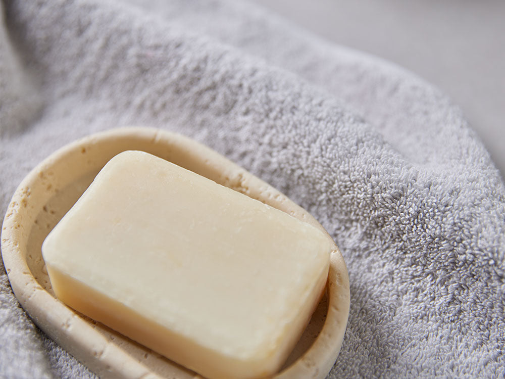 Naturseife in einer ovalen Seifenschale aus beigem Naturstein auf einem hellgrauen Baumwollhandtuch im Badezimmer.