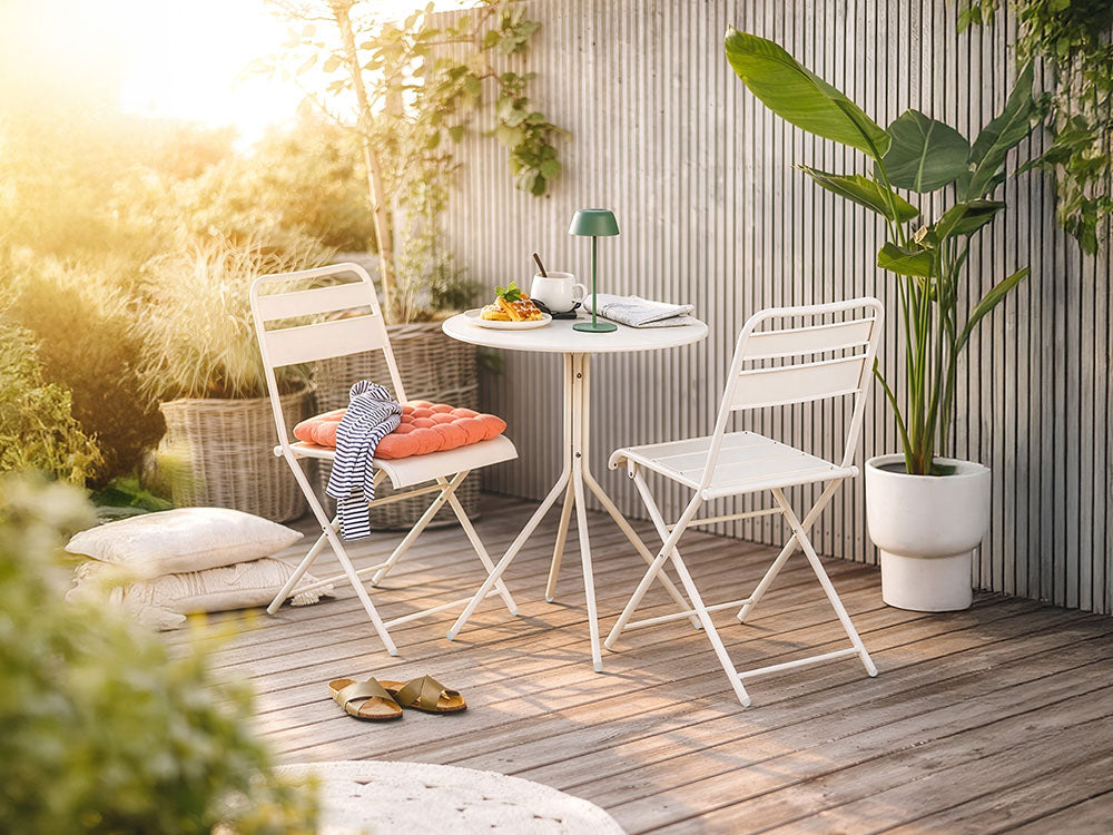 Weiße Bistro-Gartenmöbel aus Metall auf einer Holzterrasse mit grüner LED Lampe, Sitzkissen und einer großen Zimmerpflanze im Topf.