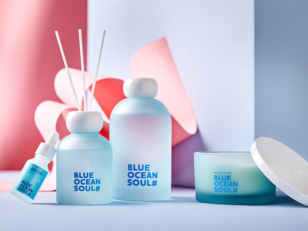 Butlers Blue Ocean Soul Set aus Duftkerze, Raumduft mit Stäbchen und ätherischem Öl in hellblauen Glasflaschen vor rosa Hintergrund.