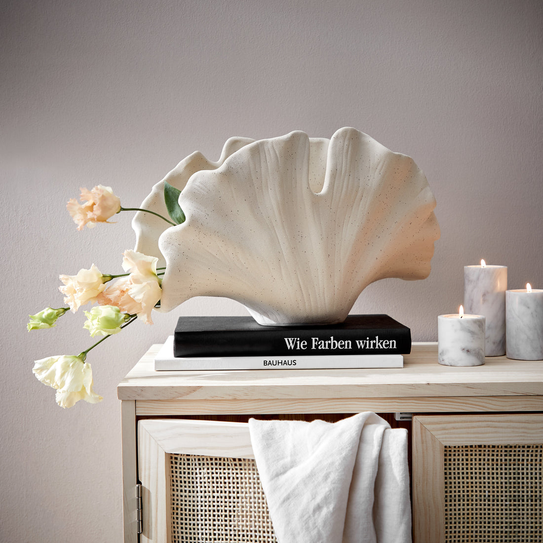 Große beige Keramikvase in organischer Fächerform auf Büchern neben Marmor-Stumpenkerzen auf einer hellen Holzkommode.
