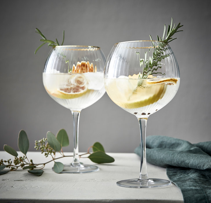 Zwei hochwertige, geriffelte Gingläser mit edlem Goldrand, einladend mit Zitrone und Gewürzen auf einem weißen Tisch angerichtet – die perfekten Cocktailgläser für deine stilvolle Hausbar.