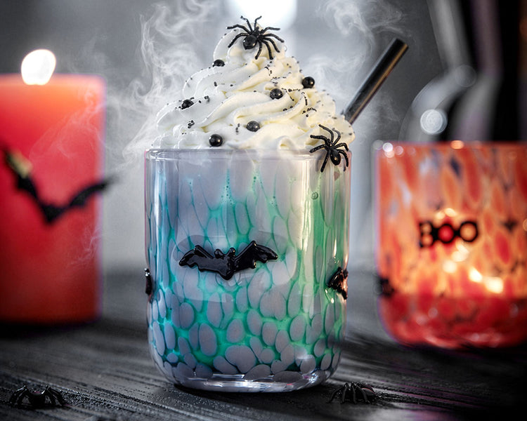 Halloween-Drink in einem türkis-gemusterten Glas mit Sahnehaube, schwarzen Zuckerperlen sowie Spinnen- und Fledermaus-Deko, umgeben von Kerzen und leichtem Nebel.