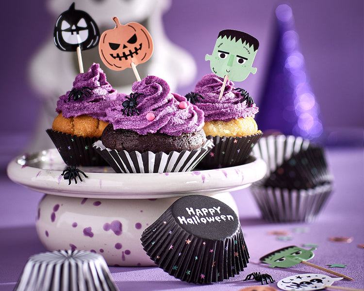 Halloween-Cupcakes mit lilafarbener Creme, Spinnen-Deko und Halloween-Steckern in Kürbis-, Geister- und Frankenstein-Optik auf einer Tortenplatte.