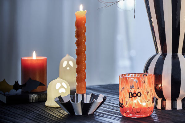 Halloween-Dekoration mit orangefarbenen Kerzen, kleinen Geisterlichtern, Fledermäusen und schwarz-weiß gestreifter Vase.