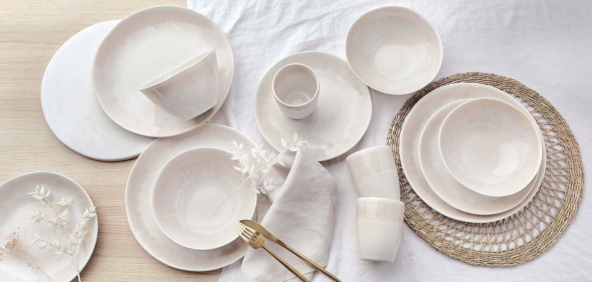 Cremeweißes Steinzeug-Geschirrset mit Tellern, Schüsseln und Bechern auf einem Jute-Tischset und weißem Leinentuch im minimalistischen Scandi-Stil.