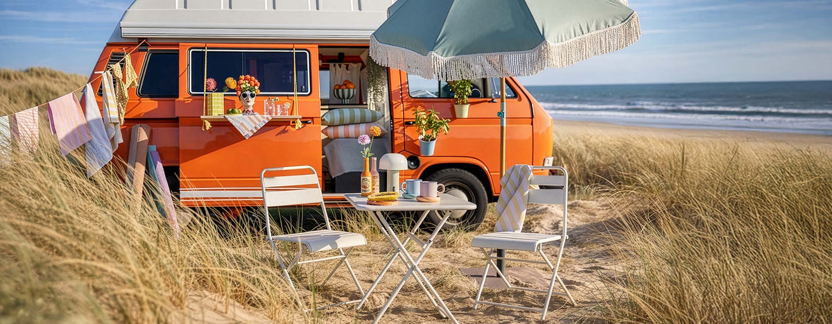 Orangefarbener Retro-Campingbus am Strand mit weißen Gartenmöbeln aus Metall, pastellfarbenem Sonnenschirm und gestreiften Outdoor-Textilien.