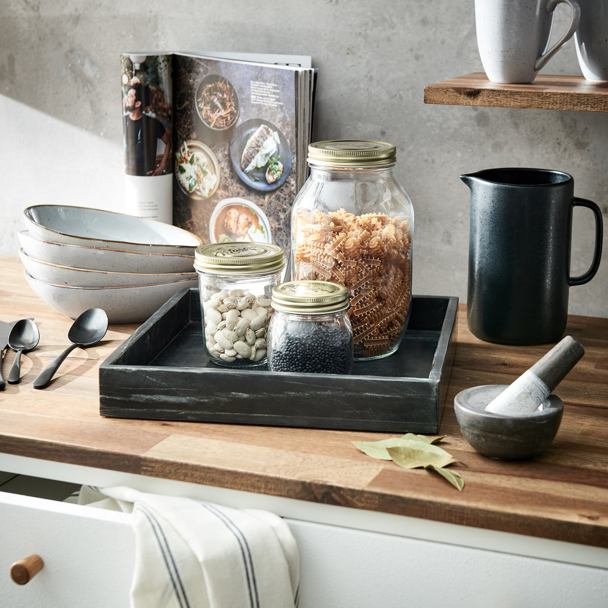 Vorratsgläser mit Golddeckeln auf Holztablett neben Mörser und Keramik