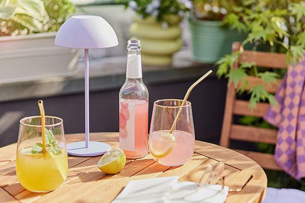 Holztisch im Freien mit rosa Kaltgetränken in Gläsern, einer fliederfarbenen LED Tischleuchte aus Metall und grünen Gartenpflanzen.