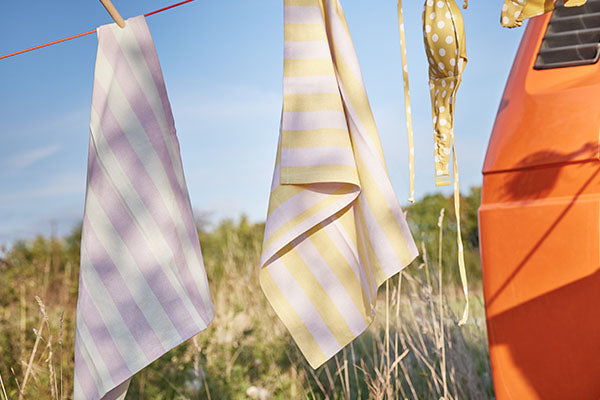 Gestreifte Geschirrtücher in Pastellgelb und Flieder hängen an einer Wäscheleine im Freien neben einem orangefarbenen Campingbus.