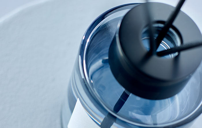 Draufsicht auf einen Raumduft-Flakon aus blauem Glas mit schwarzen Diffuser-Stäbchen für die Wohnung.