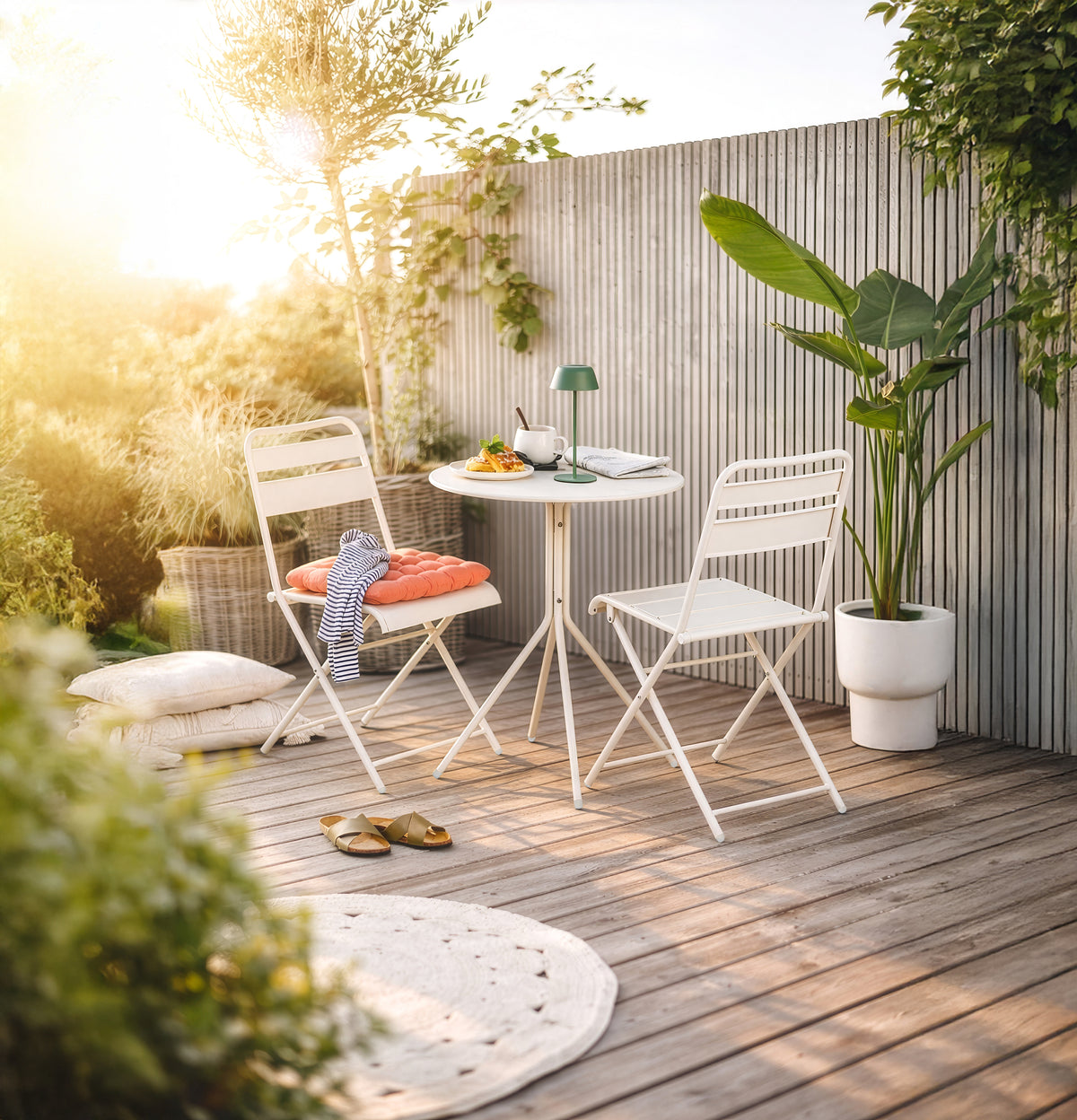 Weißes Gartenmöbel Set aus Metall mit einem runden Bistrotisch und klappbaren Stühlen auf einer Holzterrasse bei Sonnenuntergang.