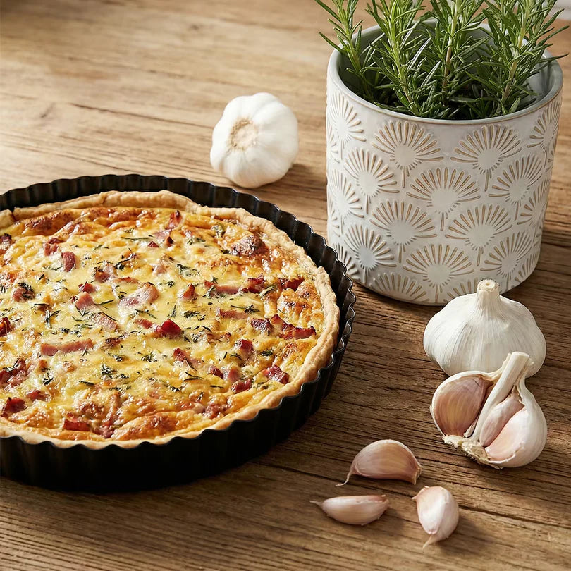 Quiche in schwarzer Tarteform neben Rosmarintopf und Knoblauch