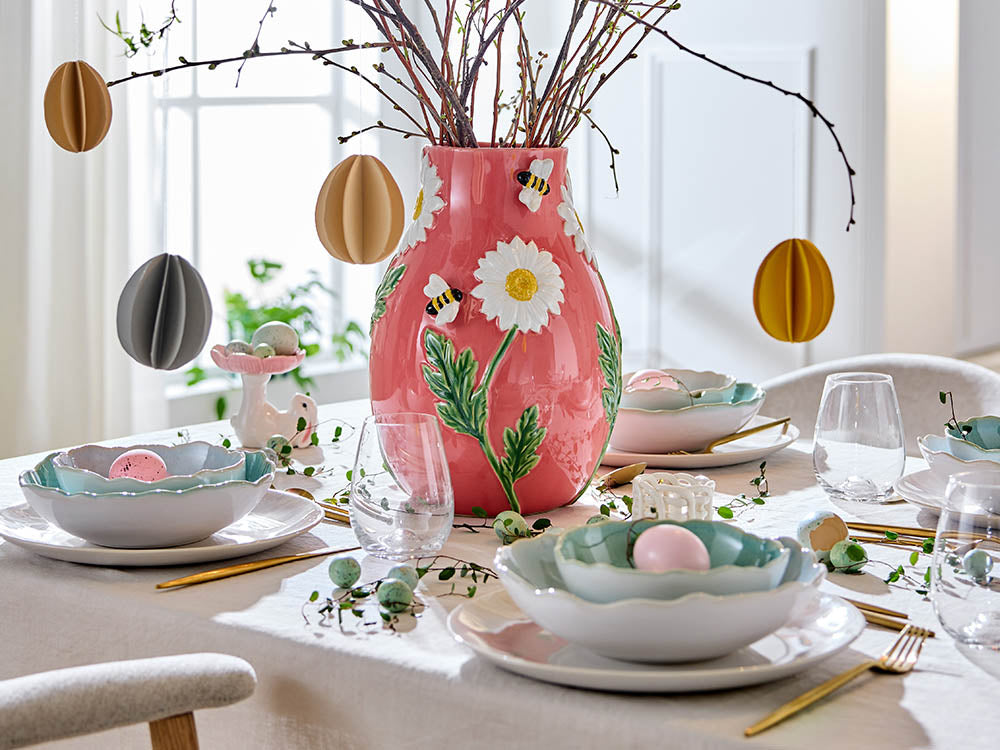 Große rosa Keramikvase mit plastischen Bienen und Gänseblümchen auf einem gedeckten Ostertisch mit pastellblauem Geschirr und hängenden Papiereiern.