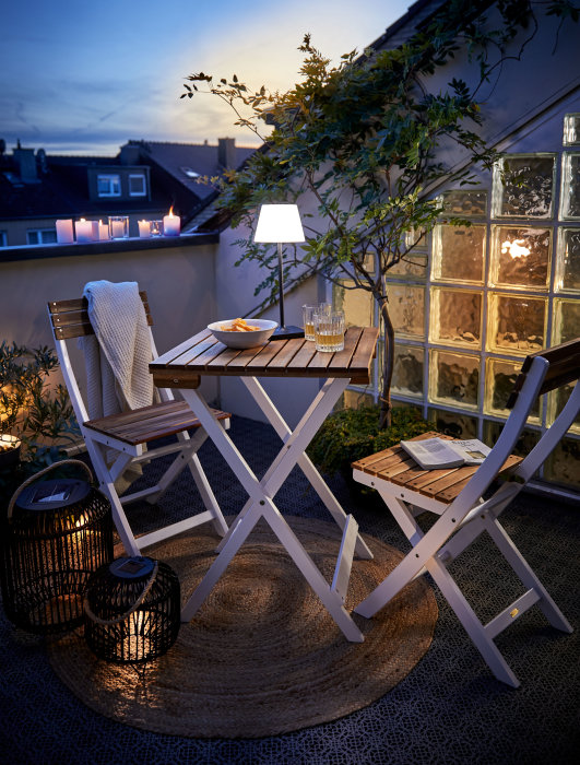 Schwarze LED-Tischleuchte auf Holztisch neben Balkonstühlen und dekorativen Windlichtern aus Metall auf dem Boden in abendlicher Atmosphäre.