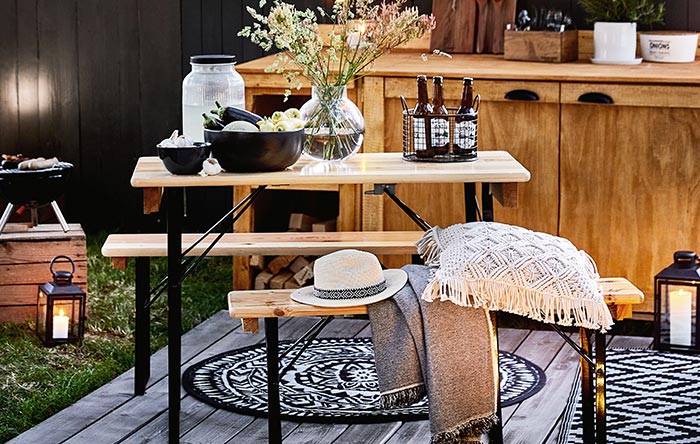 Picknickbank und Tisch aus hellem Holz auf einer Terrasse mit Laternen, Flaschenträger und Dekokissen.