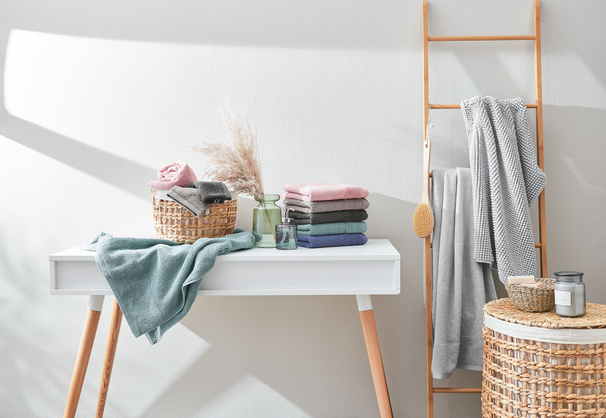 Stapel bunter Handtücher aus Baumwolle in Rosa, Grau und Blau auf weißem Tisch neben einer Holzleiter mit grauen Duschtüchern im modernen Badezimmer.