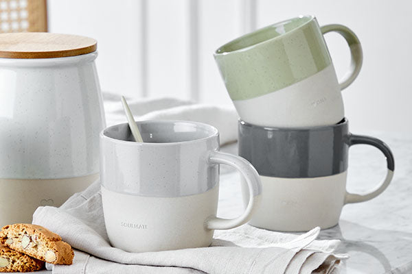 Drei stapelbare Kaffeebecher aus zweifarbigem Steingut in Grau, Grün und Cremeweiß neben einer Vorratsdose auf einer Leinenserviette.