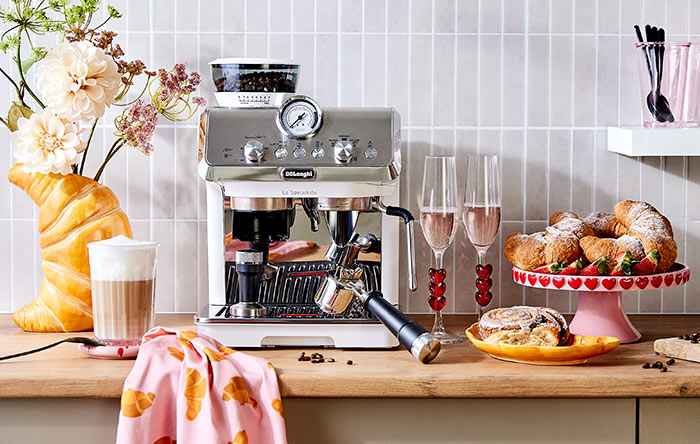 Espressomaschine aus Edelstahl mit Siebträger in einer Küche, daneben Gläser, Croissants auf einem Ständer und ein Tuch.