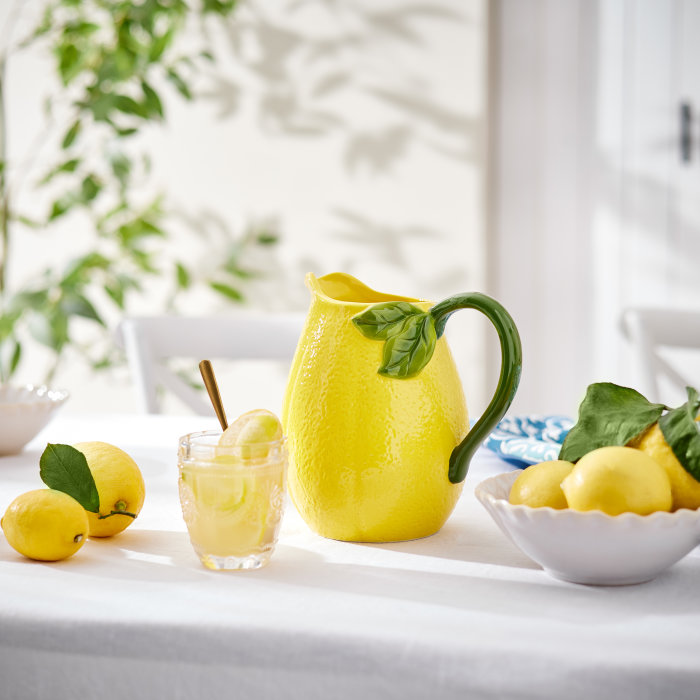 Gelber Keramikkrug in Zitronenform mit grünem Henkel auf weiß gedecktem Tisch mit frischen Zitronen und Limonadenglas.