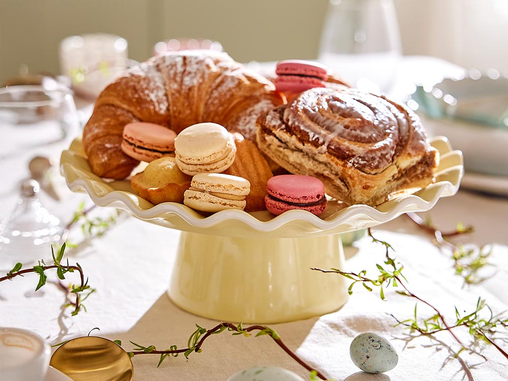 Gelbe Tortenplatte aus Keramik mit gewelltem Rand, präsentiert mit Croissants, Macarons und Gebäck auf einer hellen Ostertafel.