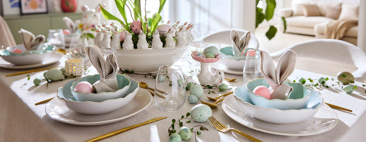 Festliche Ostertafel mit rosa Keramikvase, farbigen Ostereiern, pastellblauen Schalen und Servietten in Hasenform auf einer hellen Tischdecke.