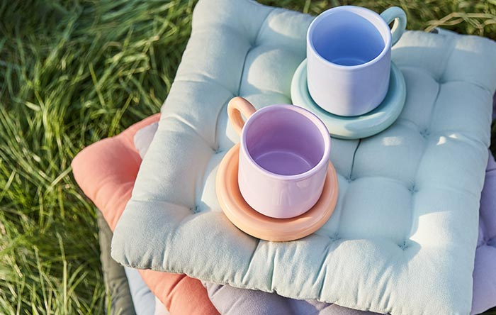 Gestapelte Textil-Sitzkissen in Pastellfarben (Blau, Rosa, Lila) auf einer grünen Wiese für draußen.