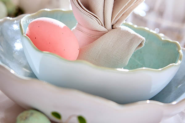 Detailaufnahme von zwei pastellblauen Steingut-Schalen mit gewelltem Rand und einer beigen Serviette in Hasenohr-Form.