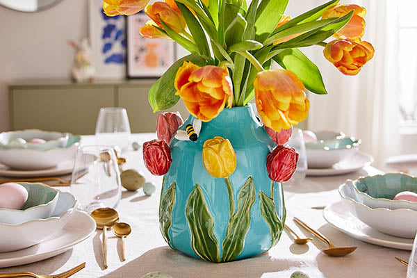 Türkisfarbene Keramikvase mit plastischen Tulpenblüten, gefüllt mit orange-gelben Tulpen auf einer weißen Tischdecke.