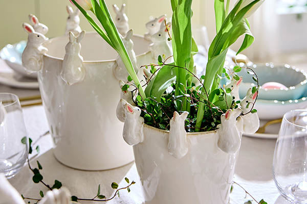 Weiße Übertöpfe aus Keramik mit umlaufenden Relief-Hasenfiguren, bepflanzt mit Frühlingsblumen auf einem gedeckten Ostertisch.