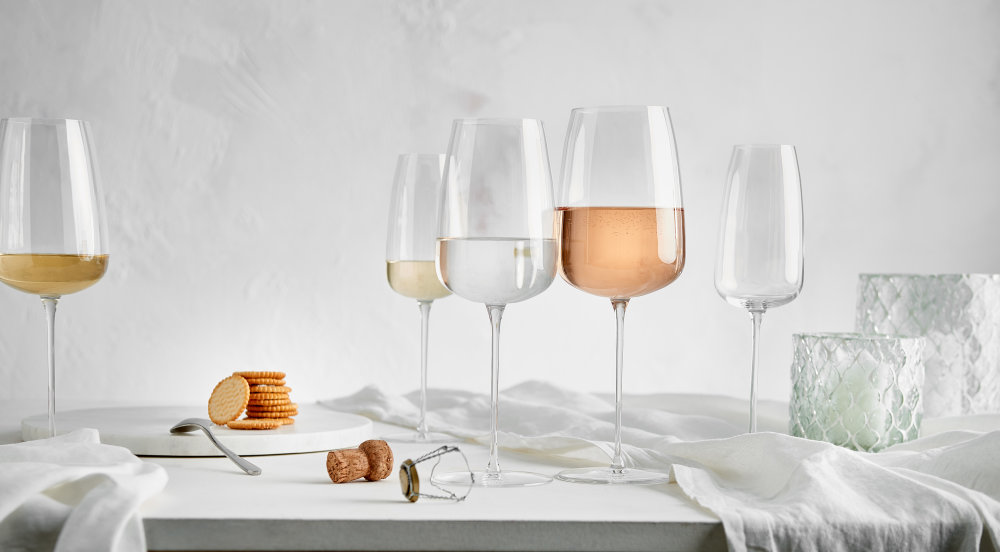 Verschiedene Weingläser aus transparentem Kristallglas mit Weißwein und Rosé auf hellem Tisch mit Textil-Dekoration im minimalistischen Stil.